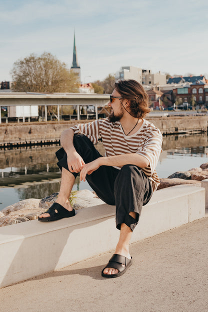 Black Miku sandals by Omaking, featuring a minimalist design, worn casually by a man by the waterfront.