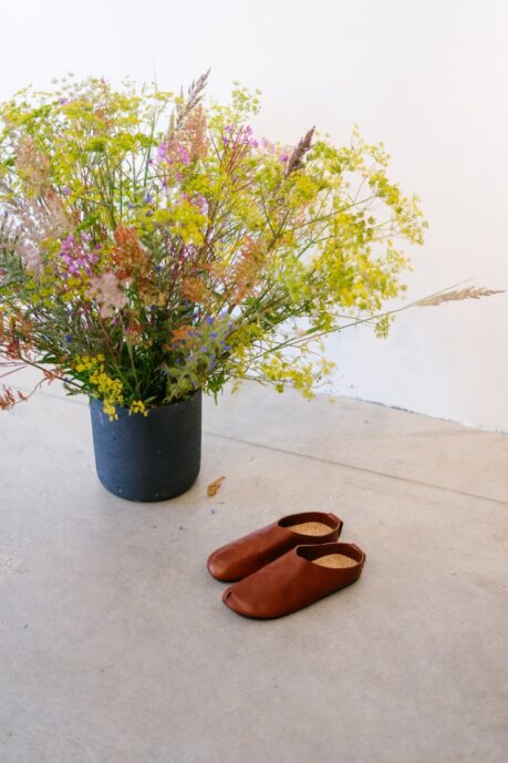 Brown Kyoto slippers by Omaking with an open toe design, placed beside a flower pot on a concrete floor.